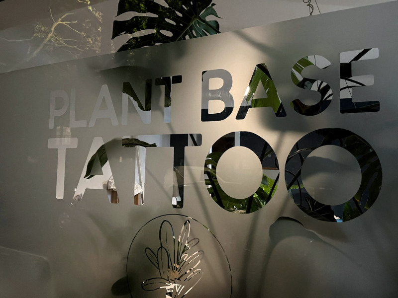 Plant Base Studio von Außen, Fenster mit der Schrift Plant Base Tattoo, dahinter Pflanzen.
